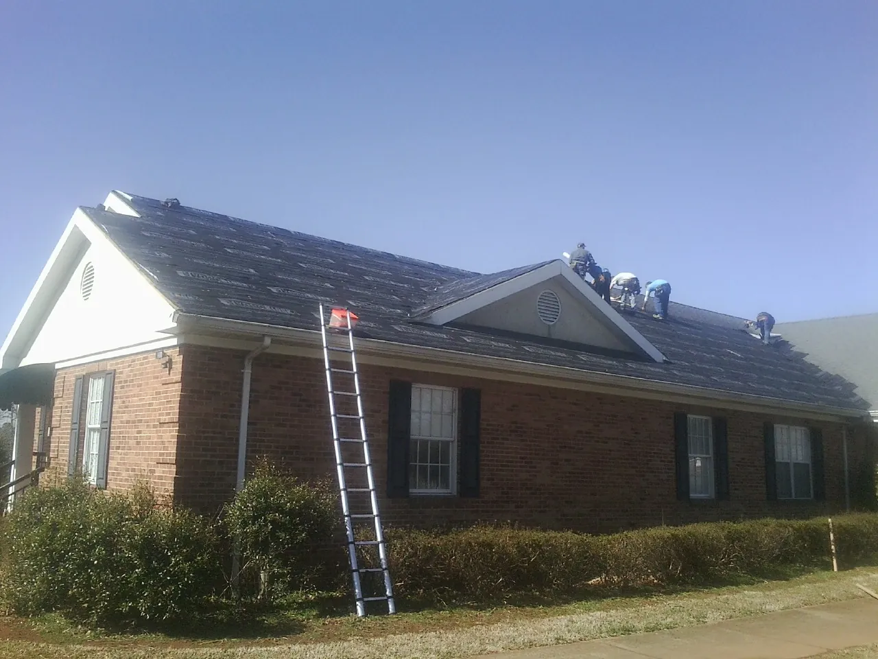 Crew tearing shingles off a brick ranch roof with a ladder set — Emergency Roof Repair work by our crew in Sundance