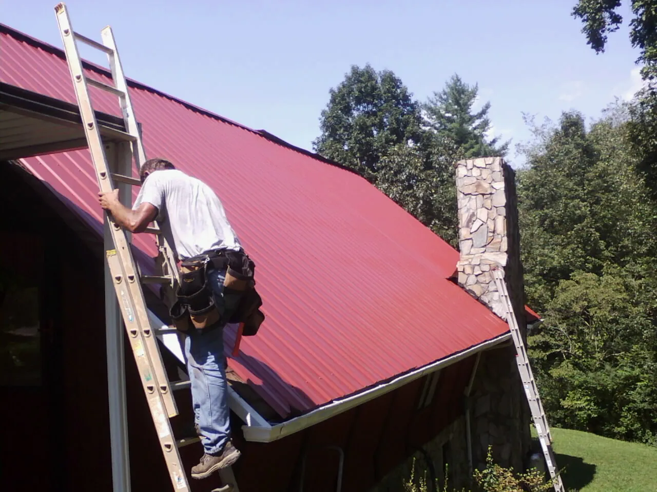 Roofer on a ladder beside a red metal roof and stone chimney — Soffit & Fascia Repair by Roofing Brothers in Sundance